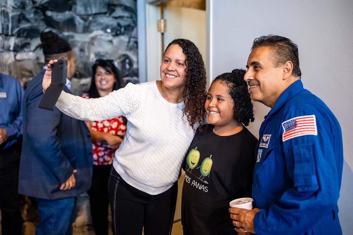 A woman takes a selfie with a child and Astronaut José Hernández, who is wearing a blue NASA flight suit. The group is smiling, and other attendees are visible in the background during a Castro event at Metropolitan State University of Denver